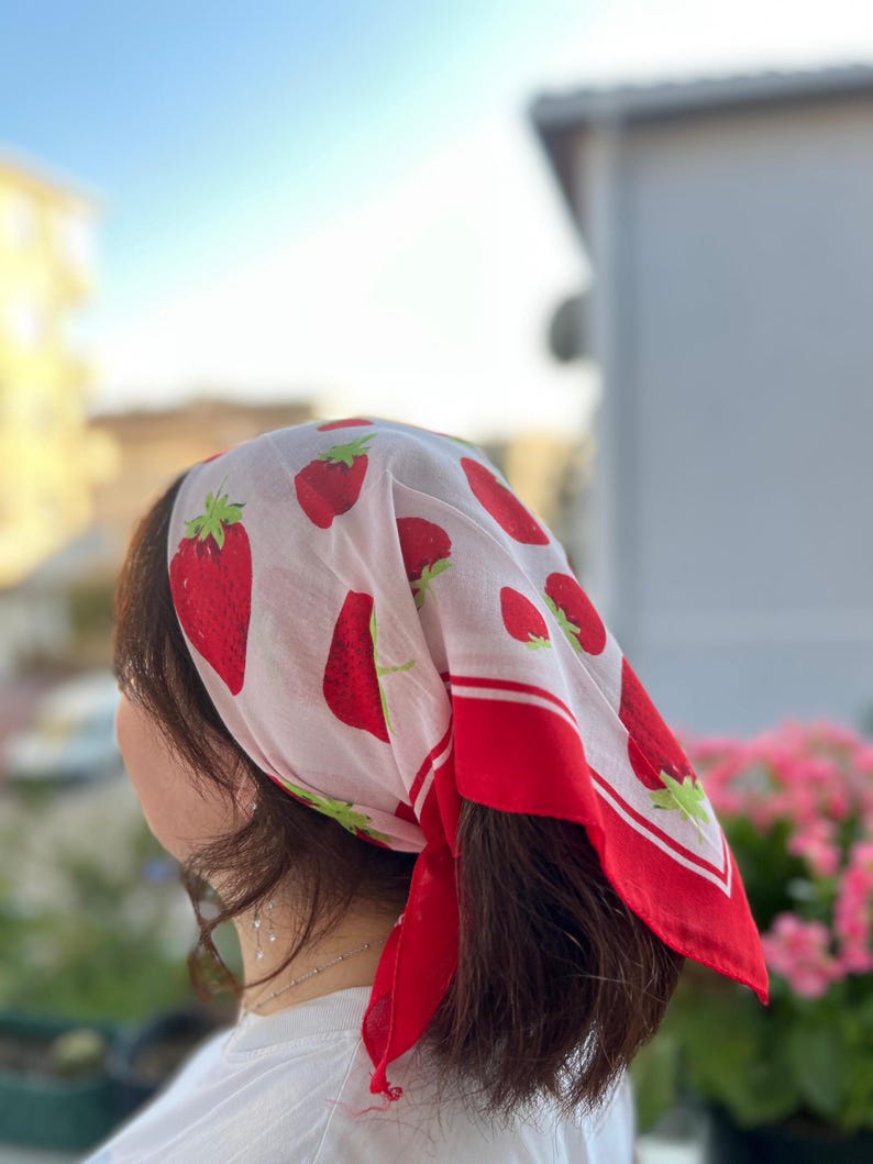 May include: A white headscarf with a red strawberry pattern and a red border. The strawberries are in various sizes and have green stems. The scarf is tied around the head.