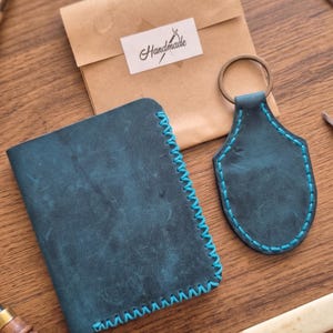 May include: A dark blue leather passport holder and a matching shield-shaped keychain, both featuring bright blue contrast stitching. A brown paper envelope with a white label reading "Handmade" is positioned behind the leather items.