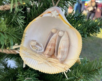 Op de afbeelding: Een kerstversiering in de vorm van een kerststal gemaakt van schelpen, met een grote schelp als stal en kleinere schelpen die Maria, Jozef en het kindje Jezus voorstellen. De versiering is versierd met stro en een kleine ster.