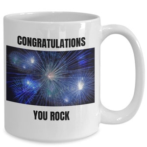 May include: White ceramic mug with a black rectangle featuring a blue and white fireworks display. The mug has the words "CONGRATULATIONS" and "YOU ROCK" printed in black.
