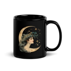May include: Black ceramic mug with a crescent moon design featuring a woman with flowing hair and a crown of stars. The background is a dark blue with gold stars.