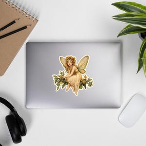 May include: A vintage illustration of a fairy with wings, sitting on a branch with yellow flowers. The fairy is wearing a white dress and a flower crown.