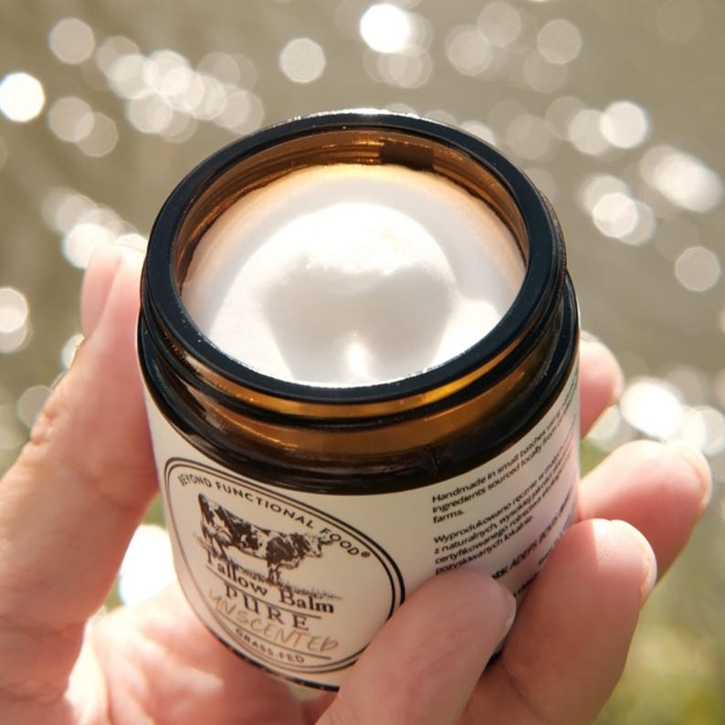 May include: An open amber glass jar containing white tallow balm. The white label features a cow illustration and the text Beyond Functional Food, Tallow Balm, Pure Unscented, Grass-Fed. The jar is held, showing the product texture inside.