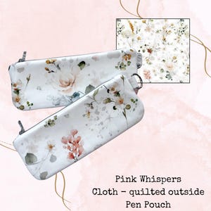 May include: Two white pen pouches with a floral design featuring pink and peach roses, green leaves, and other wildflowers. The pouches have a zip closure and a small silver ring. The text "Pink Whispers Cloth - quilted outside Pen Pouch" is visible.