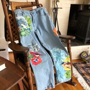 May include: Light blue denim jeans with floral and bird embroidery. The embroidery features a variety of colors, including red, yellow, blue, and green.