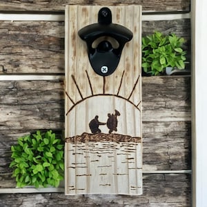 May include: Wooden bottle opener with a burnt wood design of a sunset and two figures standing on a rock. The bottle opener is mounted on a wooden board.