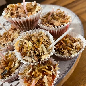 Op de afbeelding: Een close-up van een groep kleine, ronde, bruine en witte kaarsen in papieren cupcakevormpjes. De kaarsen zijn bedekt met houtsnippers en hebben een roze lont.