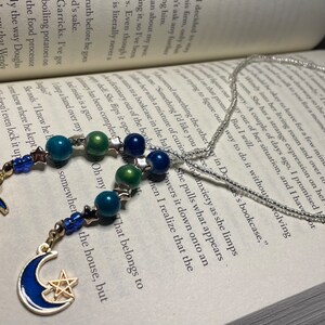 May include: Two silver beaded necklaces with crescent moon charms. The charms are blue and green with gold accents and star details. The necklaces are on a book with text.