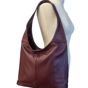 May include: A burgundy leather shoulder bag with a large, open design. The bag has a single shoulder strap and a textured surface. The bag is displayed on a white mannequin.