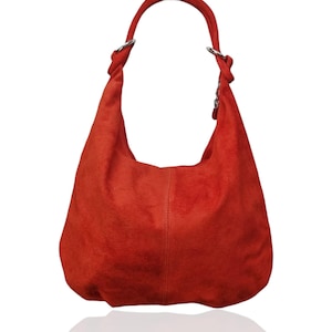 May include: A red suede hobo bag with a single shoulder strap. The bag has a curved shape and a soft texture. Silver-toned rings connect the strap to the bag. The bag is set against a white background with a subtle reflection.
