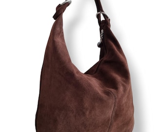 Op de afbeelding: Een bruine suède hobo-tas met een enkele schouderriem. De tas heeft zilverkleurige metalen ringen waar de riem aan vastzit en een kleine bedel. De tas heeft een zacht, nonchalant ontwerp.