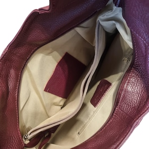 Puede incluir: Vista interior de un bolso de mano de cuero burdeos con forro de tela beige. El bolso tiene un bolsillo con cremallera y un peque&ntilde;o bolsillo deslizante.