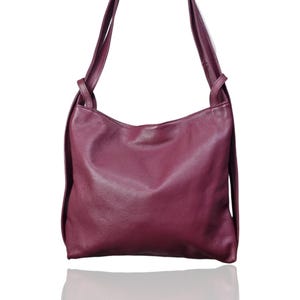 Peut inclure: Un sac à bandoulière en cuir bordeaux foncé avec une surface texturée. Le sac a deux longues anses et une forme arrondie. Le sac se reflète sur la surface blanche en dessous.