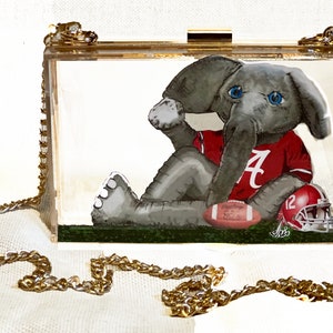 Op de afbeelding: Een transparante acryl clutch met een gouden kettingband. De tas heeft een cartoon afbeelding van een olifant die een rood shirt draagt met een witte "A" erop. De olifant houdt een voetbal en een helm vast.