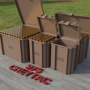 May include: Several brown storage containers, some open, filled with brass-colored ammunition. The containers are labeled "375" and the red text "CHEYTAC 375" is in the foreground. The containers are on a concrete surface with green grass in the background.