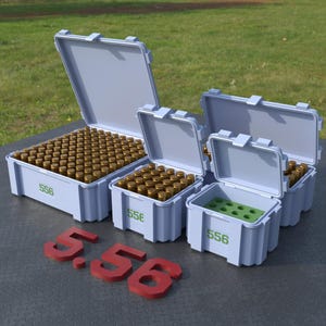 Könnte beinhalten: Mehrere hellblaue Aufbewahrungsbehälter, einige offen, gefüllt mit messingfarbener Munition. Die Behälter sind mit "556" in Grün beschriftet. Rote "5.56" Ziffern befinden sich im Vordergrund. Die Behälter stehen auf einer grauen Oberfläche mit grünem Gras im Hintergrund.