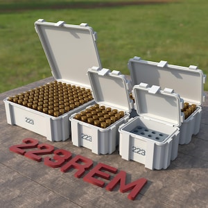 May include: Four white plastic ammo boxes with lids open, labeled "223 REM". The largest box is full of gold-colored bullets, the other three boxes are empty.