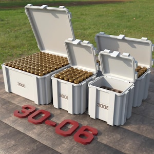 May include: Several white storage containers with open lids, filled with brass-colored ammunition. The containers are labeled "3006" and are arranged on a wooden surface. Red 3D letters spell out "30-06" in the foreground.