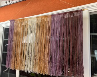Puede incluir: Un tapiz de pared hecho de hilo en tonos de blanco, beige y morado. El hilo está enhebrado en largos hilos y cuelga de una varilla de madera.