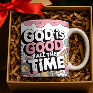 May include: White ceramic mug with the text "GOD IS GOOD ALL THE TIME" in black and pink lettering. The mug has a pink rim and is decorated with flowers, a rainbow, and other colorful designs. The mug is presented in a gift box with a red bow.