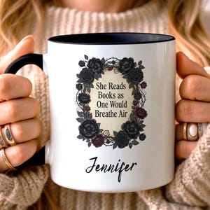 Op de afbeelding: Witte keramische mok met een zwarte binnenkant en handvat. De mok heeft een bloemenontwerp met zwarte rozen en de tekst "She Reads Books as One Would Breathe Air" en de naam "Jennifer".