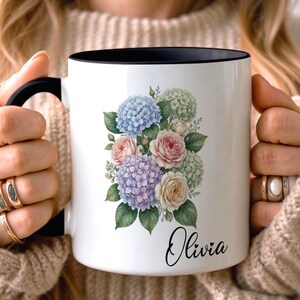 Op de afbeelding: Witte keramische mok met een zwarte binnenkant en handvat, met een bloemenontwerp van blauwe, paarse en roze bloemen met groene bladeren. De naam "Olivia" staat in een sierlijk lettertype op de mok.