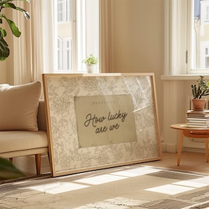 Puede incluir: Un gran estampado enmarcado con un marco de madera clara. El estampado presenta un fondo de patrón floral con un diseño de postal vintage que dice "How lucky are we". El estampado está apoyado contra una pared en una habitación bien iluminada.
