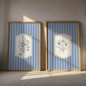 May include: Two framed botanical art prints. Each print features a gold frame, a blue and white striped background, and a central cream-colored cartouche. One print displays a detailed illustration of flowers, while the other showcases a bouquet of lilies.