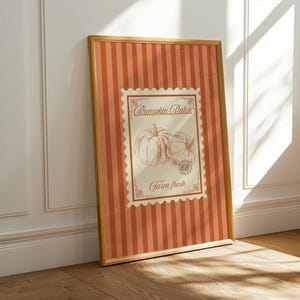 May include: A framed print with a vintage design, featuring a pumpkin illustration. The print has an orange striped background and a central stamp-like image with the words "Pumpkin Patch" and "Farm Fresh". The frame is a warm gold colour.