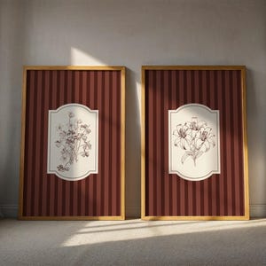 May include: Two framed botanical art prints. Each features a gold frame, a burgundy and cream striped background, and a central cream-colored cartouche with a detailed floral illustration in brown. One depicts flowers, the other lilies.