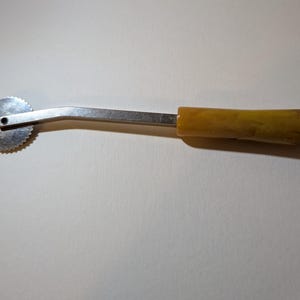 May include: A vintage pastry wheel with a circular, serrated blade and a curved metal arm. The handle is a light amber color. This kitchen tool is used for cutting dough.