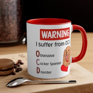 Puede incluir: Taza de cerámica blanca con borde y asa rojos. La taza tiene un cartel de advertencia que dice "ADVERTENCIA Sufro de TOC... Trastorno obsesivo del cocker spaniel". Un cocker spaniel de dibujos animados con un corazón rojo está representado en la taza.