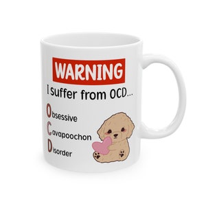 Op de afbeelding: Witte keramische mok met een handvat. De mok heeft de tekst "WARNING I suffer from OCD..." met het acroniem OCD uitgeschreven. Een cartoon Cavapoochon hond met een roze hart staat ook op de mok.