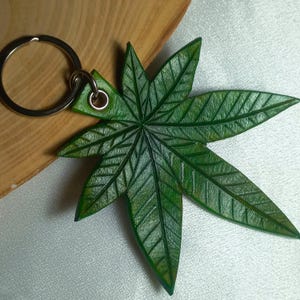 Op de afbeelding: Een groene leren sleutelhanger in de vorm van een marihuanablad. Het blad heeft gedetailleerde nerven en een gestructureerd oppervlak.