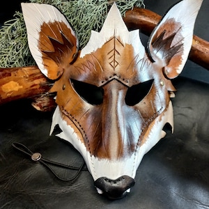 Può includere: Una maschera di volpe in pelle marrone e bianca con occhi neri e naso nero. La maschera ha un bordo di pelliccia bianca intorno ai bordi e un cinturino in pelle per fissarla alla testa.