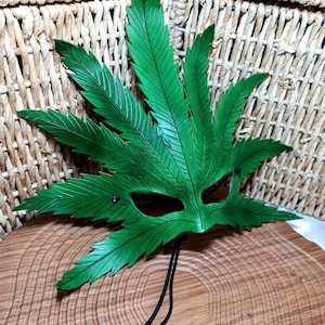 Op de afbeelding: Een groen leren masker in de vorm van een marihuanablad. Het masker heeft ooggaten en een zwart touwtje om het vast te binden.