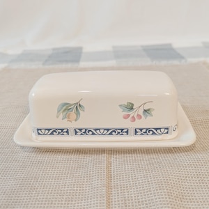 May include: Cream-colored rectangular butter dish with a lid. The dish features a blue decorative border and fruit illustrations. The lid has a matching design. The butter dish is on a white rectangular base.