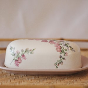 May include: A ceramic butter dish with a lid, featuring a floral design in pink and green on a cream background. The dish sits on a matching pink plate, perfect for kitchen decor.