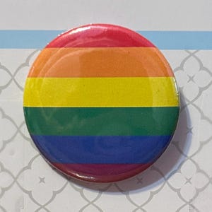 May include: A circular pin-back button featuring a horizontal rainbow flag design. The button displays six distinct stripes in red, orange, yellow, green, blue, and purple, arranged in a stacked pattern with a smooth, glossy finish across the surface.