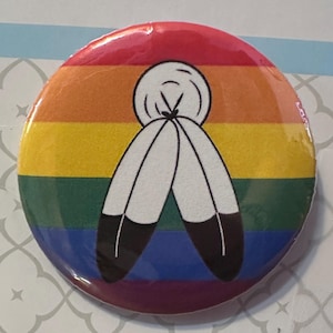 May include: A circular pin-back button featuring a horizontal rainbow stripe background in red, orange, yellow, green, blue, and purple. Centered on the button is a graphic illustration of two white feathers with black tips tied together at the top.