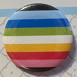 May include: A circular pin-back button featuring a series of horizontal stripes in light blue, dark blue, lime green, white, yellow, red, and pink. The button has a glossy finish and a black border visible at the top and bottom edges of the design.