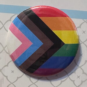 May include: A circular pin-back button featuring the Progress Pride flag design. The graphic includes a white, pink, light blue, brown, and black chevron on the left side, with horizontal red, orange, yellow, green, blue, and purple stripes.