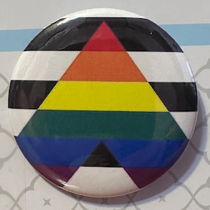 May include: A circular pin-back button featuring a central triangle design composed of horizontal stripes in red, orange, yellow, green, blue, and purple. The background of the button consists of alternating horizontal black and white stripes.