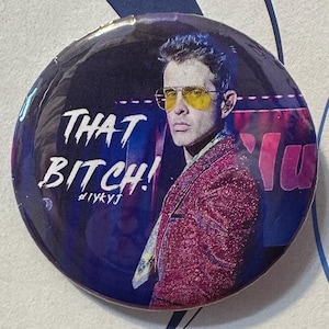 Könnte beinhalten: Runder Button mit dem Porträt eines Mannes mit gelb getönten Gläsern und einem roten Blazer. Der Button trägt den Text "THAT BITCH!" in weißer, stilisierter Schrift. Der Hashtag #iykyj ist ebenfalls sichtbar.