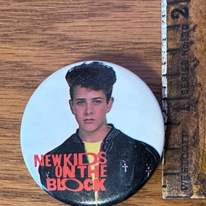 Puede incluir: Un botón redondo con un retrato de un joven y el texto "NEW KIDS ON THE BLOCK" en rojo. El hombre lleva una chaqueta negra sobre una camisa amarilla. Una regla de madera está al lado del botón.