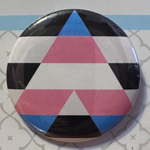 Pode incluir: Um broche redondo com um design geométrico composto por triângulos e faixas horizontais. A paleta de cores inclui preto, branco, rosa claro e azul claro, dispostos num padrão simétrico que cria uma forma triangular central.