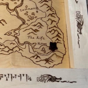 May include: A light-colored wooden board featuring a wood-burned map with geographical labels including Eastmarch, The Rift, and Riften. The border displays intricate knotwork dragon designs and runic text, all rendered in dark brown burnt lines.