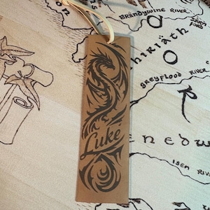 May include: A brown leather bookmark with a black dragon design and the name "Luke" in a decorative font. The bookmark has a cream-colored ribbon attached at the top. The background features a map.