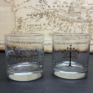 May include: Two clear glass tumblers. One features etched text in a script, the other a tree design. The background includes a map with text. The tumblers are likely themed around a fantasy book or movie.
