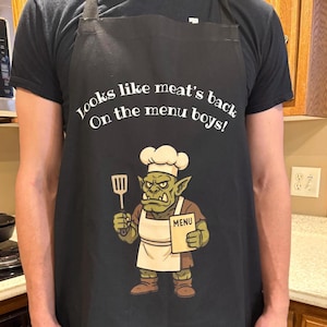 May include: Black apron with the text "Looks like meat's back on the menu boys!" and a cartoon orc chef holding a spatula and menu. The orc is wearing a chef's hat and apron.